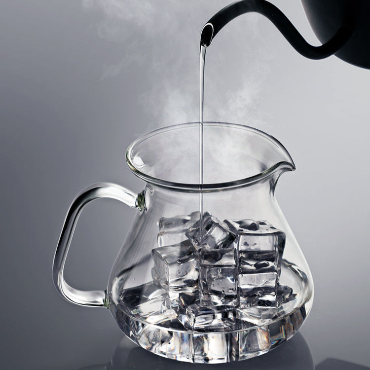 Clear glass pitcher with ice cubes being filled with hot water from a black kettle on a gray background