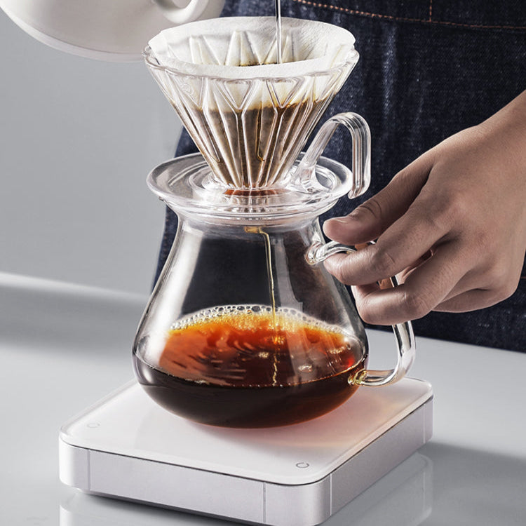 Person making coffee using a glass pour-over coffee maker on a white surface.