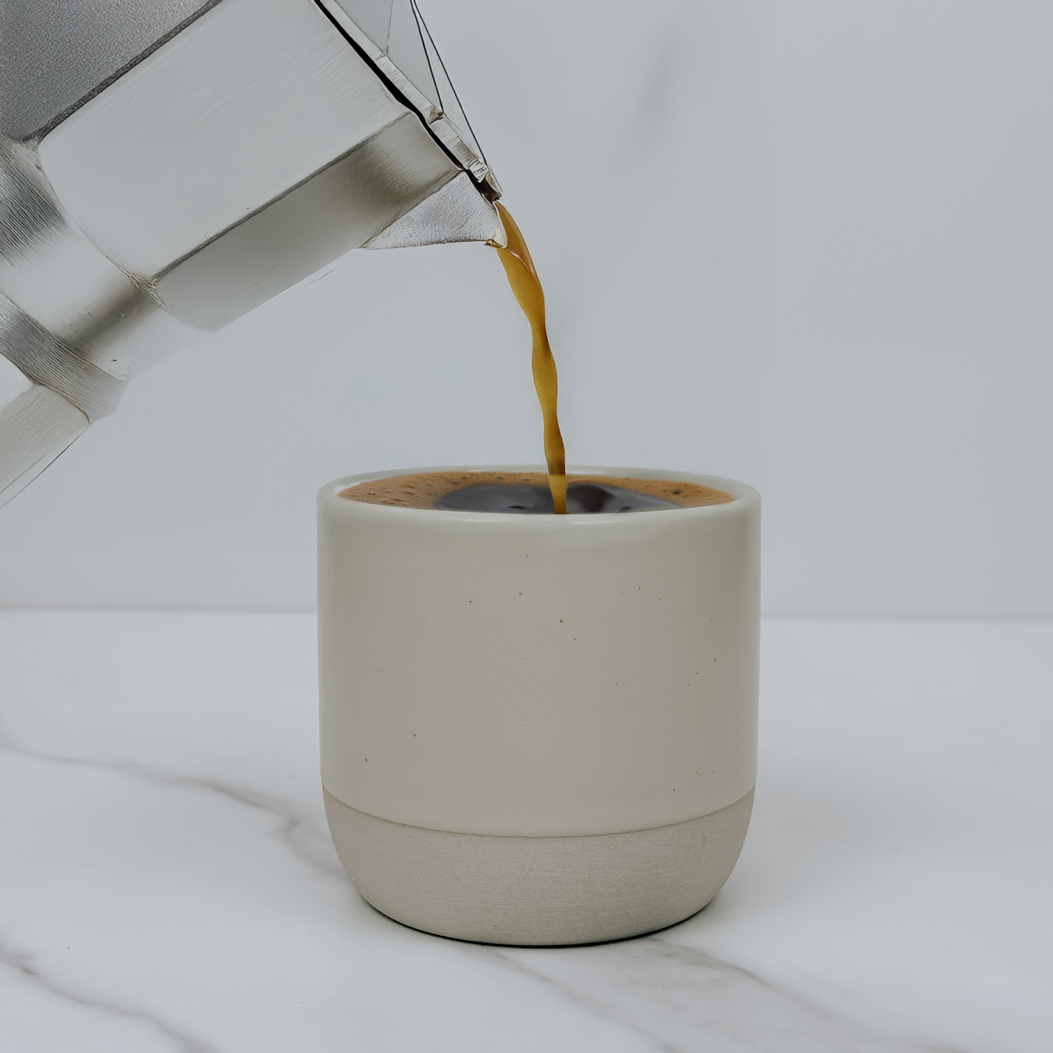 Coffee being poured from a silver kettle into a beige ceramic cup on a white background