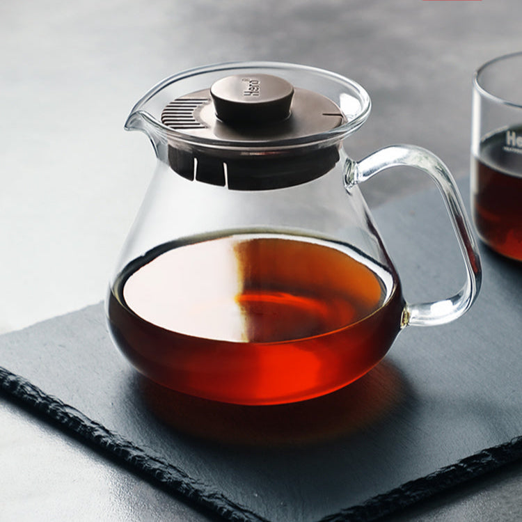 Clear glass teapot with a dark lid filled with tea on a gray surface.