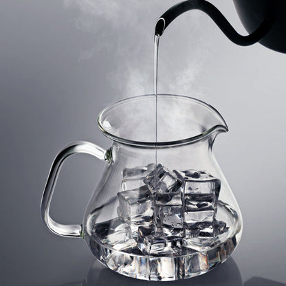 Clear glass pitcher with ice cubes being filled with hot water from a black kettle on a gray background