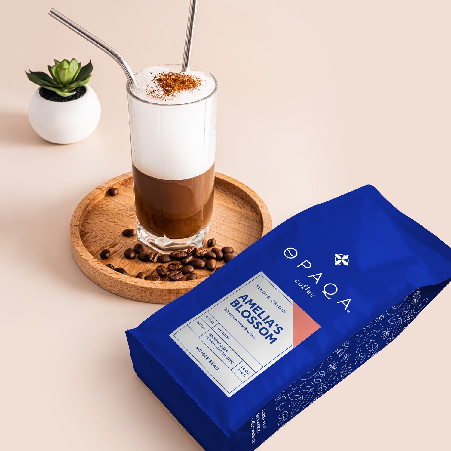 Opaqa Coffee's Amelia's Blossom coffee bag displayed next to a glass of frothy coffee topped with cinnamon. The scene includes coffee beans scattered on a wooden tray and a small plant, highlighting the sensory experience of enjoying coffee