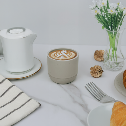 Cappuccino in a white cup with latte art, croissant, and flowers on a white table.